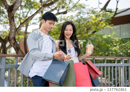 Asian couple a man showing shopping bag to woman holding coffee while standing on footbridge in park Asian couple a man showing shopping bag to woman holding coffee while standing on footbridge in park 137762861