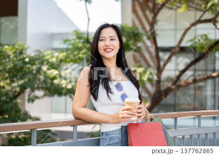 Pretty asian woman carrying shopping bags holding coffee while standing leaning on railing in park. Pretty asian woman carrying shopping bags holding coffee while standing leaning on railing in park. 137762865