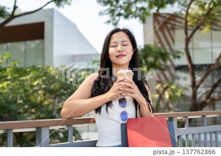 Pretty asian woman closing eyes smelling coffee as carrying shopping bags leaning on railing in park 137762866