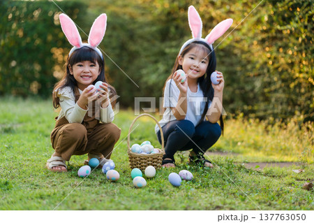 two happy child girl wearing bunny ears playing together and hunting for Easter eggs into basket on green grass in garden 137763050