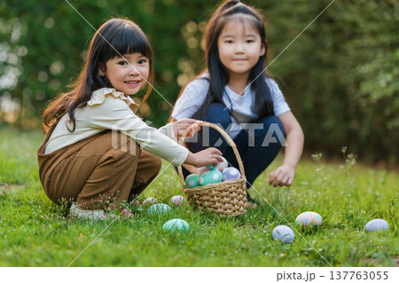 two happy child girl playing together and hunting for Easter eggs into basket on green grass in garden 137763055