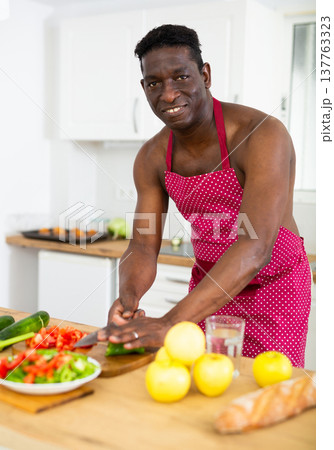 Cheerful man wearing only red apron preparing vegetable salad in home kitchen 137763323