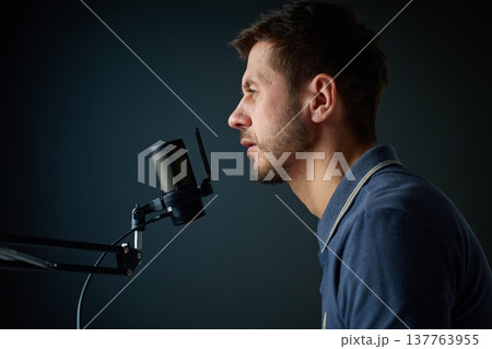 Man speaking into studio microphone against dark background Man speaking into studio microphone against dark background 137763955
