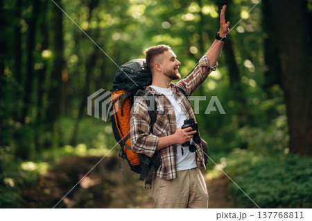Side view, standing, holding binoculars. Male tourist is with backpack in the forest Side view, standing, holding binoculars. Male tourist is with backpack in the forest 137768811