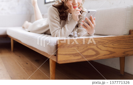 ソファに腹ばいになりスマホを使う女性 137771326