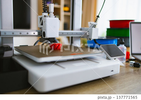 A 3D printer is actively creating a small red object on its build plate, with spools of filament visible in the background A 3D printer is actively creating a small red object on its build plate, with spools of filament visible in the background 137771565