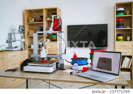 A modern 3D printer is actively creating a red object alongside spools of filament and a laptop displaying the design 137771594
