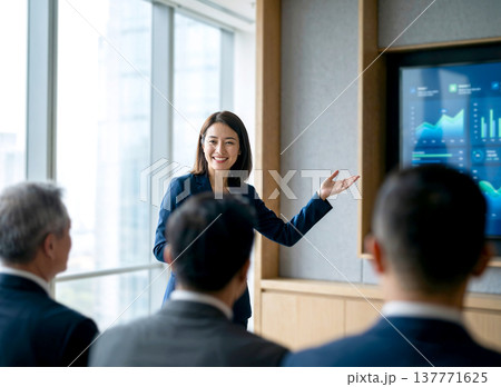 Confident business woman in a navy suit giving a presentation with data charts on a screen during a corporate meeting in a bright modern office. 137771625