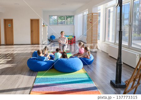Teacher reading aloud to children sitting on blue beanbags during a bright classroom story time session Teacher reading aloud to children sitting on blue beanbags during a bright classroom story time session 137773315