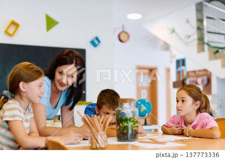 Teacher guiding elementary students as they observe a classroom terrarium and work on a science activity together 137773336