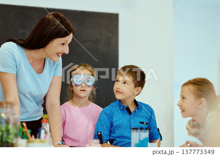 Teacher guiding curious children during a hands-on classroom science experiment with safety goggles and test tubes Teacher guiding curious children during a hands-on classroom science experiment with safety goggles and test tubes 137773349