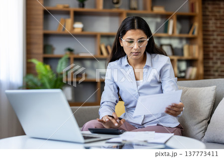 Young woman wearing glasses sitting on a sofa. Managing personal finances. Reviewing documents. Using a calculator. And working on her laptop in a contemporary living room. Focusing on bills and taxes Young woman wearing glasses sitting on a sofa. Managing personal finances. Reviewing documents. Using a calculator. And working on her laptop in a contemporary living room. Focusing on bills and taxes 137773411