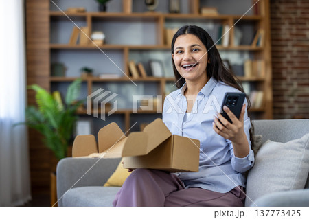 Happy young woman sitting on sofa and holding mobile phone, feeling excited after receiving her online shopping order, unboxing cardboard box with a cheerful expression 137773425