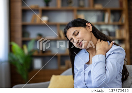 Young woman grimacing indoors, rubbing sore neck and shoulder from fatigue and poor posture, showing stress, muscle tension and discomfort while sitting at home 137773436