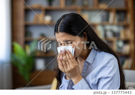 Woman feeling unwell suffering from common cold, flu or seasonal allergies, using a white tissue to blow her nose or cover a sneeze while staying home 137773443