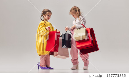 Two little girls standing together looking inside shopping bags while wearing oversized fashionable clothes Two little girls standing together looking inside shopping bags while wearing oversized fashionable clothes 137773886