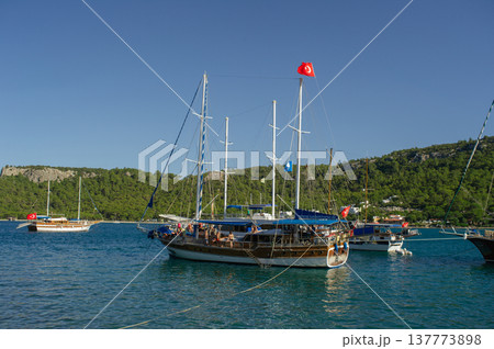 A wide horizontal shot of a large traditional wooden gulet yacht with multiple masts and a Turkish flag, anchored in a turquoise sea bay surrounded by lush green mountains under a clear blue sky. A wide horizontal shot of a large traditional wooden gulet yacht with multiple masts and a Turkish flag, anchored in a turquoise sea bay surrounded by lush green mountains under a clear blue sky. 137773898