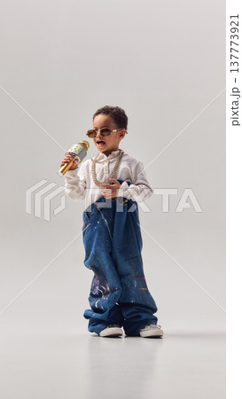 Little boy singing into microphone while wearing oversized denim jeans in studio. 137773921