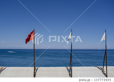 A wide horizontal shot of the Turkish national flag and two white flags flying on poles against a clear blue sky and calm sea at a seaside resort. 137773935