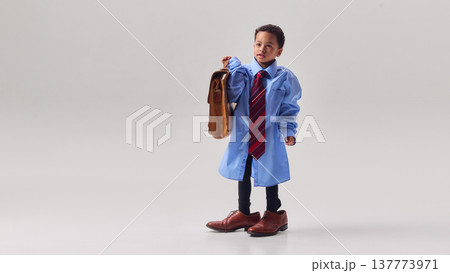 Little boy wearing oversized shirt and tie and holding large briefcase in studio. Little boy wearing oversized shirt and tie and holding large briefcase in studio. 137773971