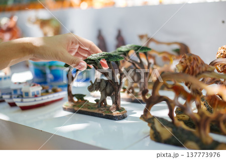 A close-up horizontal shot of a person's hand reaching for a small, detailed elephant figurine placed under a stylized wooden tree canopy, with other animal statues blurred in the background. 137773976