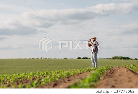 Black binoculars is in hands. Looking far away. Man farmer is on the agricultural field Black binoculars is in hands. Looking far away. Man farmer is on the agricultural field 137775062
