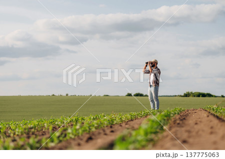 Black binoculars is in hands. Looking far away. Man farmer is on the agricultural field 137775063