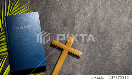 Top-down view of a blue Holy Bible, a wooden cross, and green palm branches on a dark textured stone background with copy space. Ideal for religious, Easter, or Palm Sunday themes 137775516
