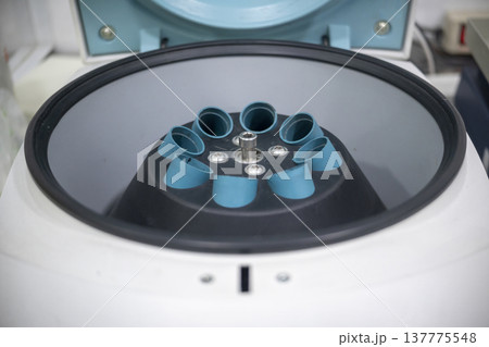 Close-up of a scientific laboratory centrifuge with its lid open, revealing the internal rotor and multiple blue tube holders ready for sample processing in a medical or research setting Close-up of a scientific laboratory centrifuge with its lid open, revealing the internal rotor and multiple blue tube holders ready for sample processing in a medical or research setting 137775548