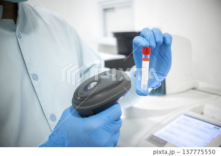 A laboratory technician wearing blue nitrile gloves scans a test tube with a handheld barcode scanner, focusing on sample tracking and diagnostic testing in a modern medical clinic 137775550