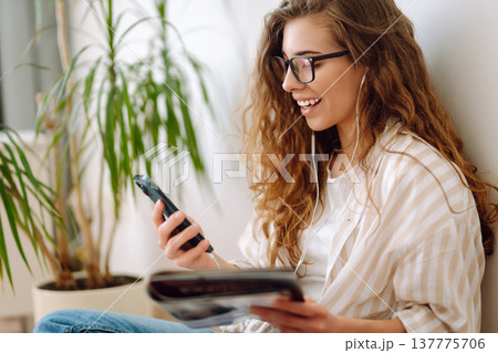 Attractive woman sits on floor in apartment with book on headphones while being distracted by phone. 137775706