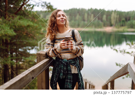 A young woman in a cozy sweater walking near a beautiful lake, smiling and using her phone 137775797