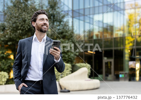Happy young businessman walking outside a modern office building, holding a smartphone and looking away with a smile, representing success, technology, and urban professional life Happy young businessman walking outside a modern office building, holding a smartphone and looking away with a smile, representing success, technology, and urban professional life 137776432