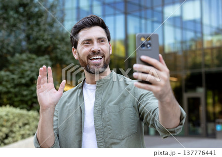 Smiling man engaging in a video call on his smartphone, waving and greeting someone virtually while standing in front of a modern building in an urban setting Smiling man engaging in a video call on his smartphone, waving and greeting someone virtually while standing in front of a modern building in an urban setting 137776441