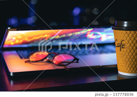 A laptop half closed in the dark with colourful glow, cup of coffee and glasses. A laptop half closed in the dark with colourful glow, cup of coffee and glasses. 137778036