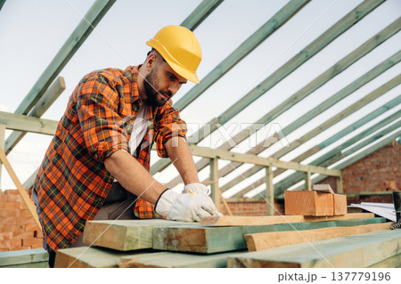 Pencil is in hand, marking place on plank. Construction worker is on the roof of a house Pencil is in hand, marking place on plank. Construction worker is on the roof of a house 137779196