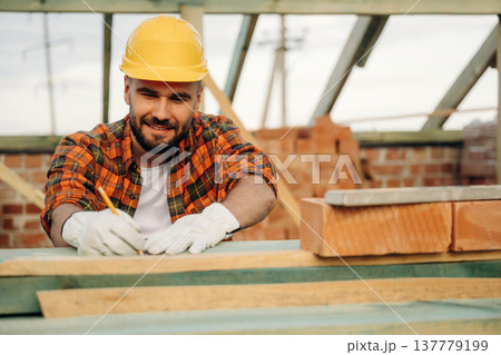 Pencil is in hand, marking place on plank. Construction worker is on the roof of a house 137779199