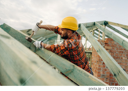 Hammer in hand, standing high up. Construction worker is on the roof of a house Hammer in hand, standing high up. Construction worker is on the roof of a house 137779203