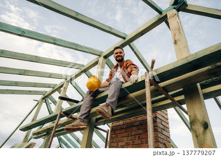 Friendly man is sitting and taking a break. Construction worker is on the roof of a house 137779207