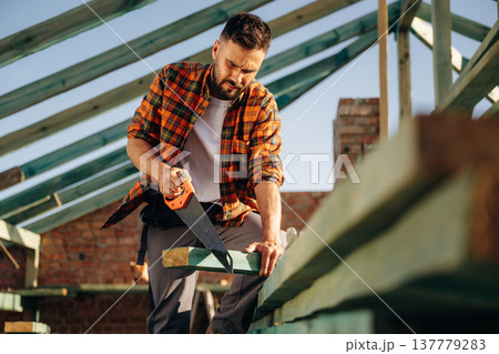 Focused, holding saw and cutting the plank. Construction worker is on the roof of a house 137779283