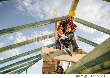 Looks down, cutting the plank with chainsaw. Construction worker is on the roof of a house Looks down, cutting the plank with chainsaw. Construction worker is on the roof of a house 137779318
