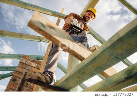 Doing the job, cutting the plank using chainsaw. Construction worker is on the roof of a house Doing the job, cutting the plank using chainsaw. Construction worker is on the roof of a house 137779327