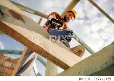 Doing the job, cutting the plank using chainsaw. Construction worker is on the roof of a house Doing the job, cutting the plank using chainsaw. Construction worker is on the roof of a house 137779329