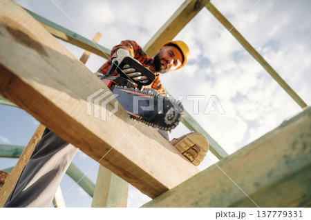 Doing the job, cutting the plank using chainsaw. Construction worker is on the roof of a house Doing the job, cutting the plank using chainsaw. Construction worker is on the roof of a house 137779331