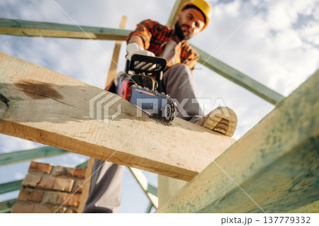 Serious facial expression, cutting the plank. Construction worker is on the roof of a house Serious facial expression, cutting the plank. Construction worker is on the roof of a house 137779332