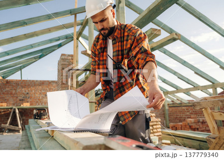 Plan of the building in hands. Construction worker is on the roof of a house Plan of the building in hands. Construction worker is on the roof of a house 137779340