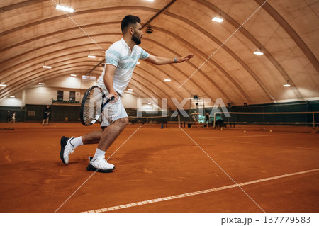 Running with racket, active time spending. Man is playing tennis indoors in the hangar Running with racket, active time spending. Man is playing tennis indoors in the hangar 137779583