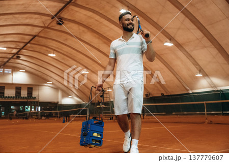 Walking forward, holding package with balls. Man is playing tennis indoors in the hangar Walking forward, holding package with balls. Man is playing tennis indoors in the hangar 137779607