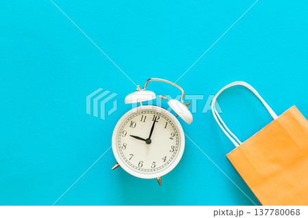 Paper bag and alarm clock on blue background, shopping time concept. Paper bag and alarm clock on blue background, shopping time concept. 137780068