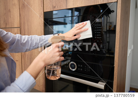 Woman wipes glass door of built in microwave oven while holding spray bottle in kitchen cabinet 137780559